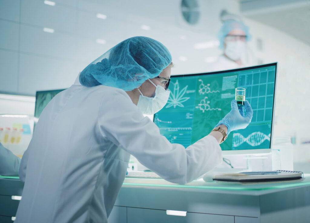 Person in a lab coat and mask examining a vial of cannabis oil in front of a computer in a laboratory.