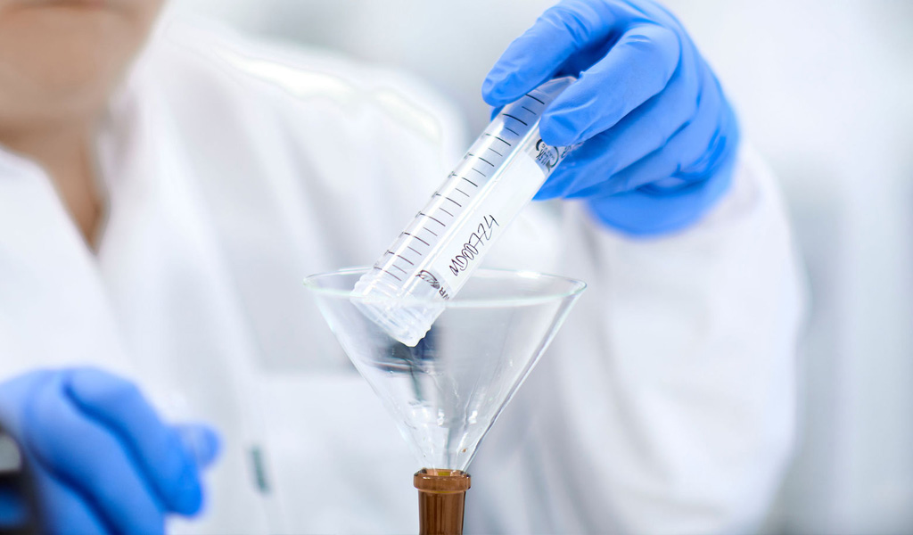 Person in a lab coat and blue latex gloves pours a test tube of cannabis oil through a funnel into a bottle.